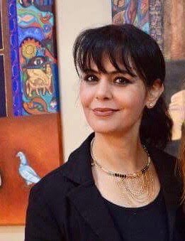 A woman stands in front of her art smiling. Her dark hair is tied back with bangs swept across her forehead. She is wearing a black blazer and shirt and silver necklace for the event.
